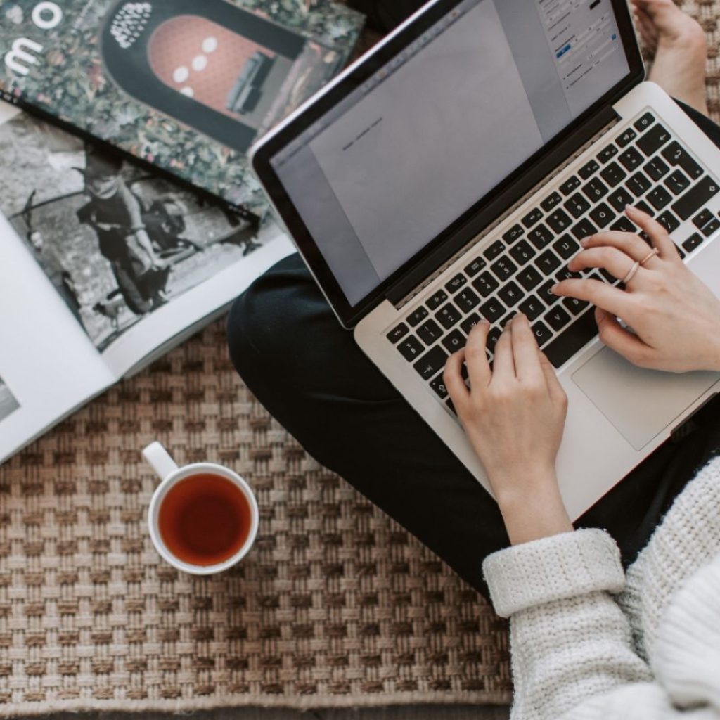 Person typing on a laptop with a cup of tea and magazines nearby.