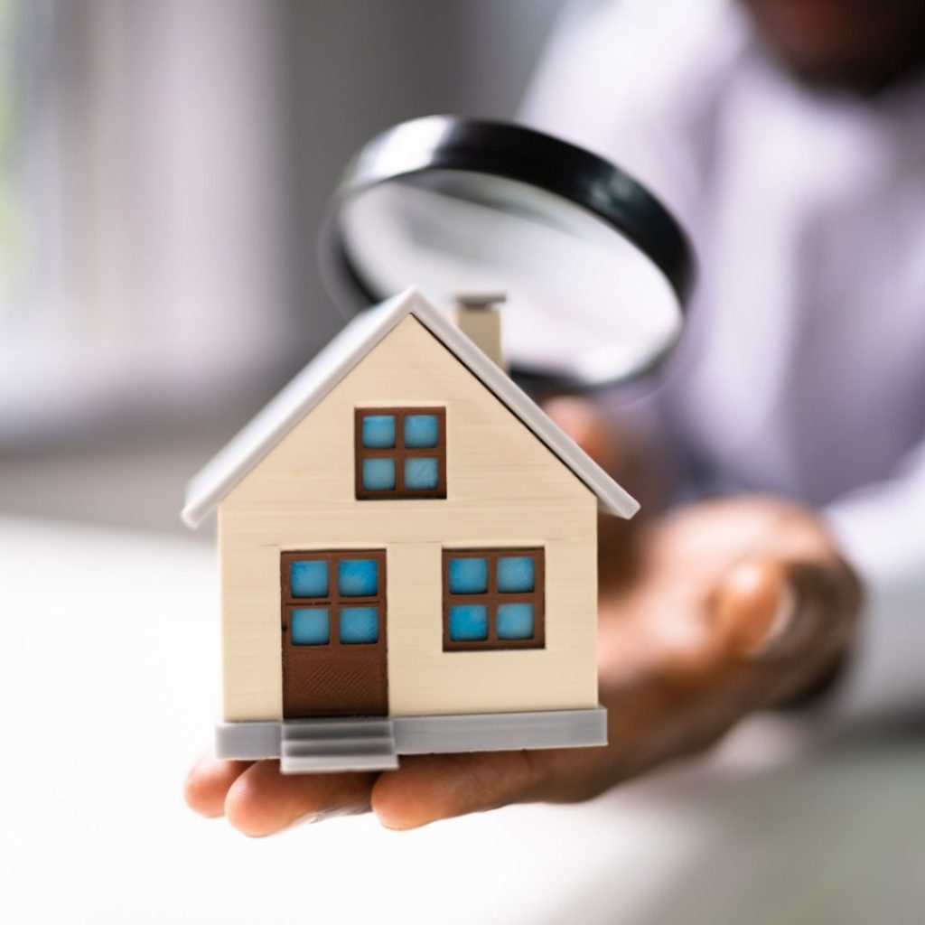 Person holding a house model with a magnifying glass inspecting it.