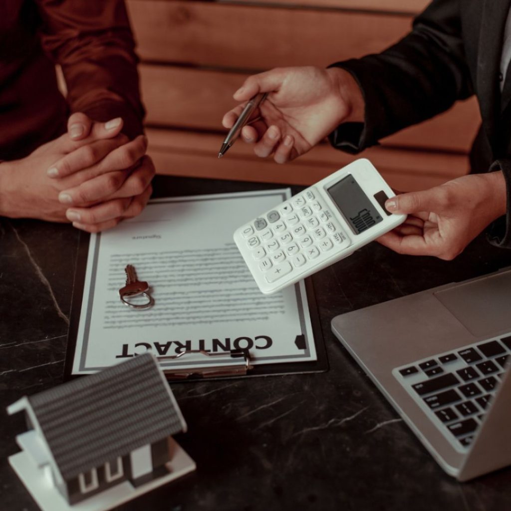 Hands discussing a real estate contract with a calculator and house model on the table.