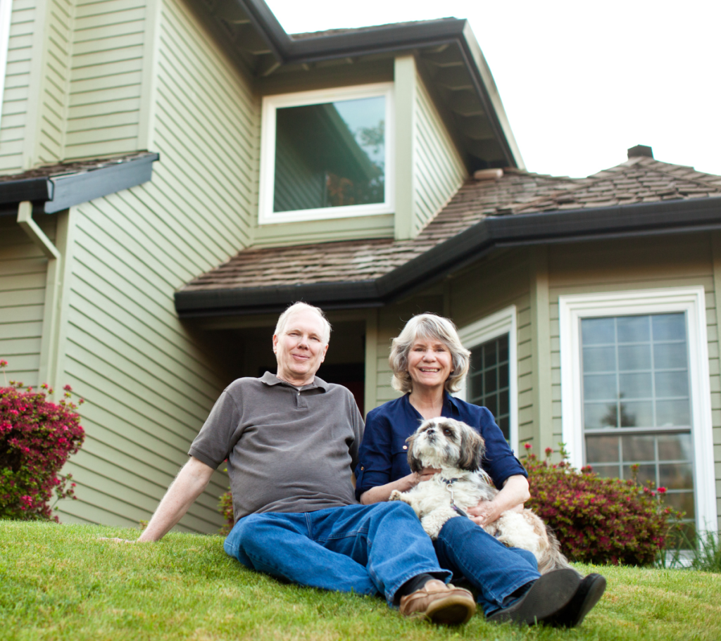 Smiling senior couple sitting on the lawn with their dog in front of their house.
