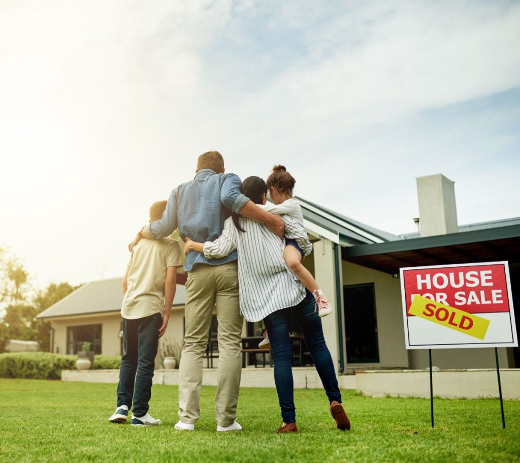 Happy family standing in front of their new home with a sold sign on the lawn.