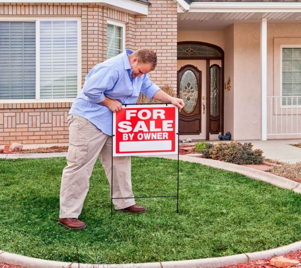 Man placing a For Sale By Owner sign in front yard of a house.