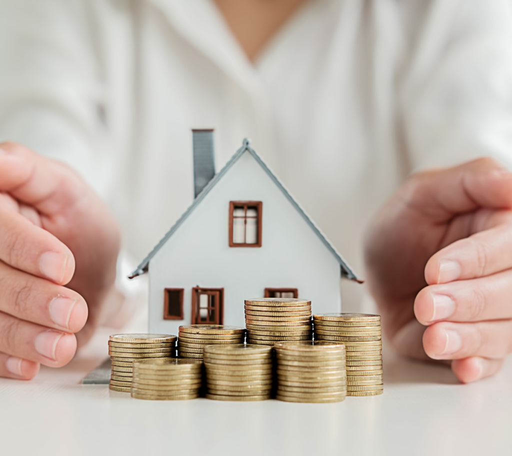 Stacked coins in front of a model house, with hands surrounding it protectively.
