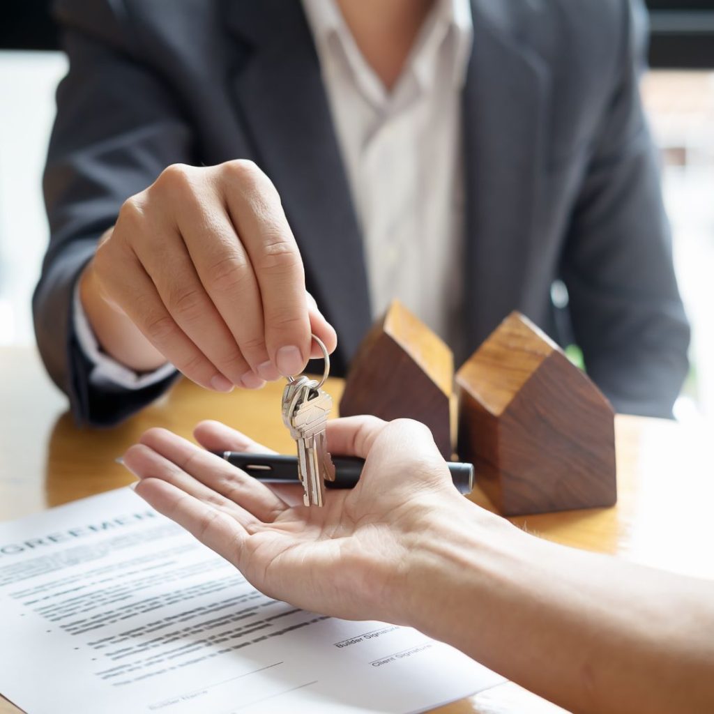 A person in a suit handing over house keys to another person across a table with a signed agreement, pen, and small wooden house models, symbolizing the completion of a real estate transaction.
