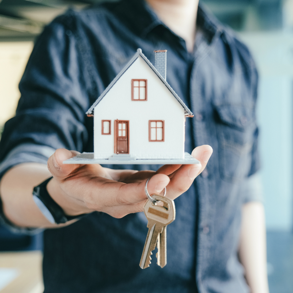 Person holding a small model house and a set of house keys, symbolizing home ownership or real estate transactions.