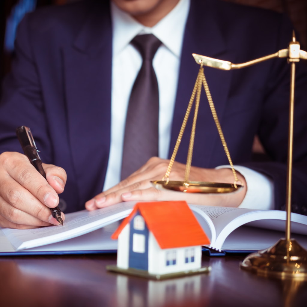 Man in suit signing documents with a model house and justice scale on desk.
