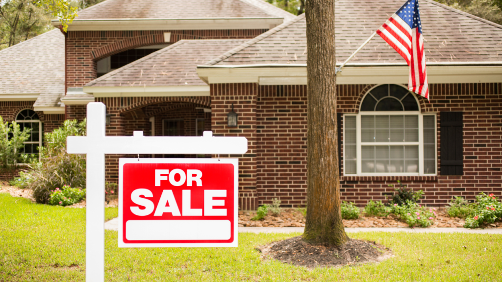 For Sale sign in front of a brick house with an American flag