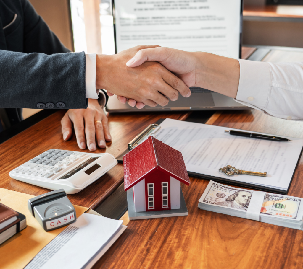 Handshake over real estate documents with house model and cash on table.
