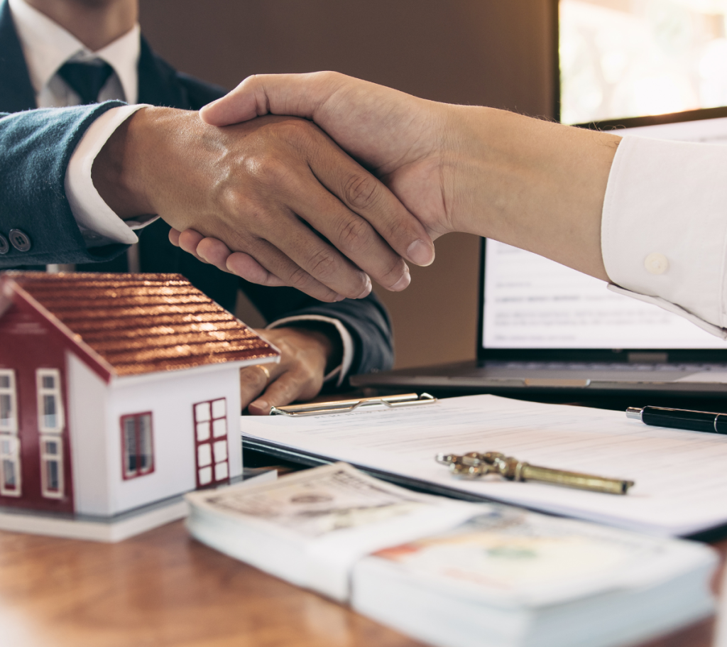 Two people shaking hands over a real estate contract with a miniature house model, keys, and cash on the table, symbolizing a successful property transaction.