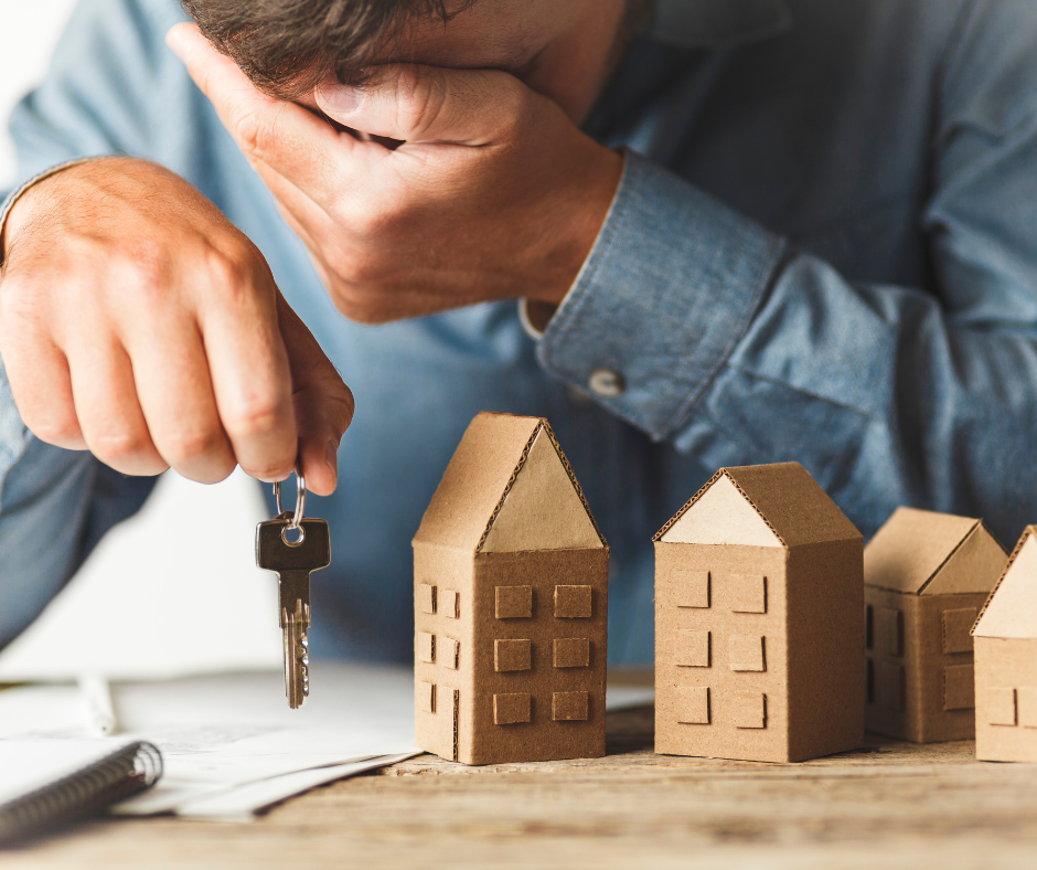 tressed man holding house keys while sitting at a desk with small cardboard house models, representing challenges and difficulties in real estate or homeownership.