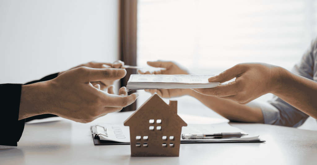 Close-up of two people exchanging money and signing documents with a small wooden house model on the table, symbolizing a real estate transaction.