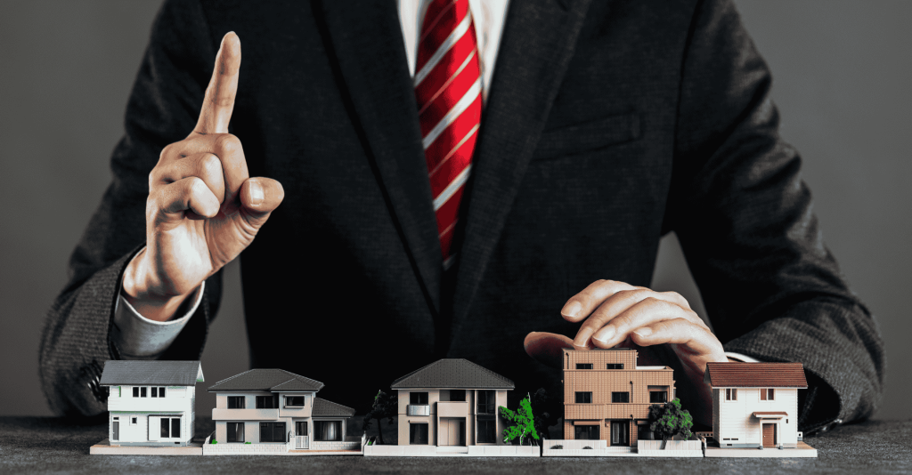 Real estate agent in a suit pointing upward while presenting miniature model houses lined up on a table.
